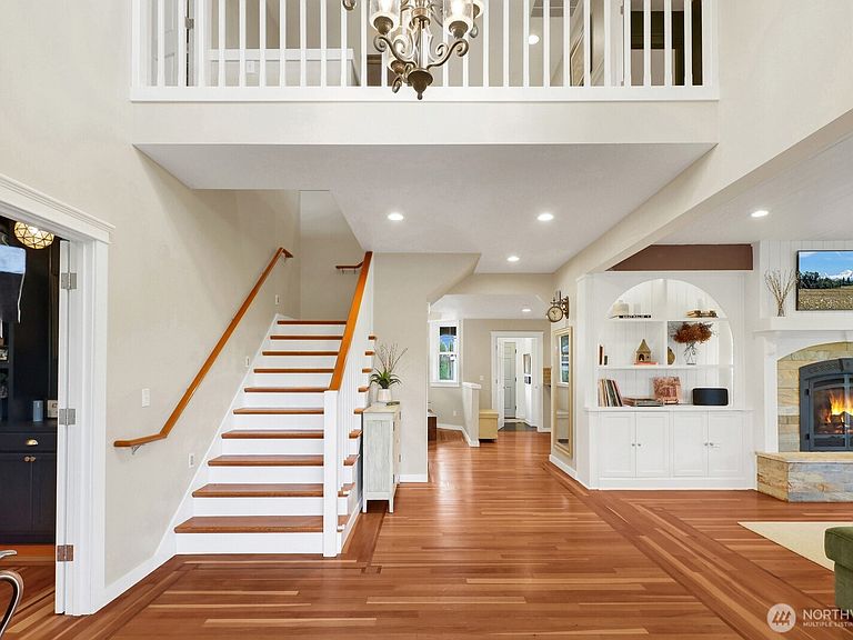 A two-story foyer with a hardwood staircase, wrought iron chandelier, and a sightline into the great room with arched built-in shelving and a stone fireplace.